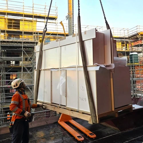 Builder in orange safety vests and helmet guiding a Superior Kitchen kitchen on a pallet that has been lifted by a crane to a floor of an under construction apartment building.